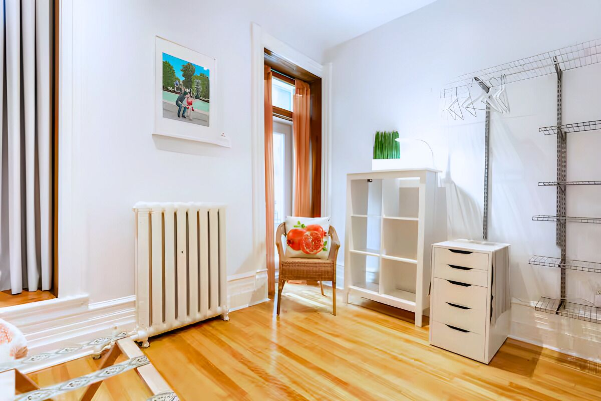 A room with a wooden floor, a radiator, a chair with a cushion, a white cabinet, and a drying rack.