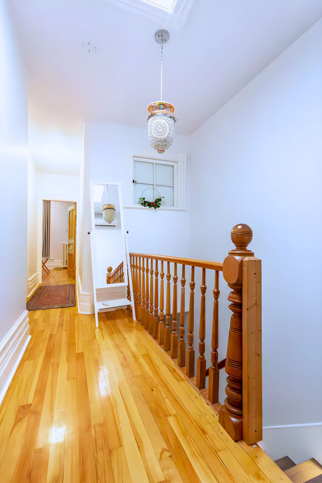 A hallway with wooden flooring, a white door, a staircase with a wooden railing, and a hanging light fixture.