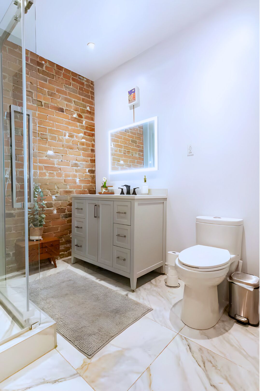 A bathroom with a brick wall, a white vanity, a toilet, and a glass shower enclosure.