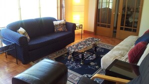 Een woonkamer met een blauwe bank, een zwarte leren poef, een glazen salontafel en een tapijt met een patroon.