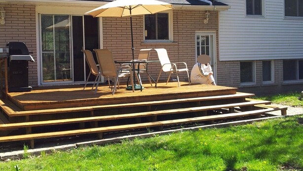 Een overdekt terras met buitenmeubilair, een barbecue en uitzicht op een huis.