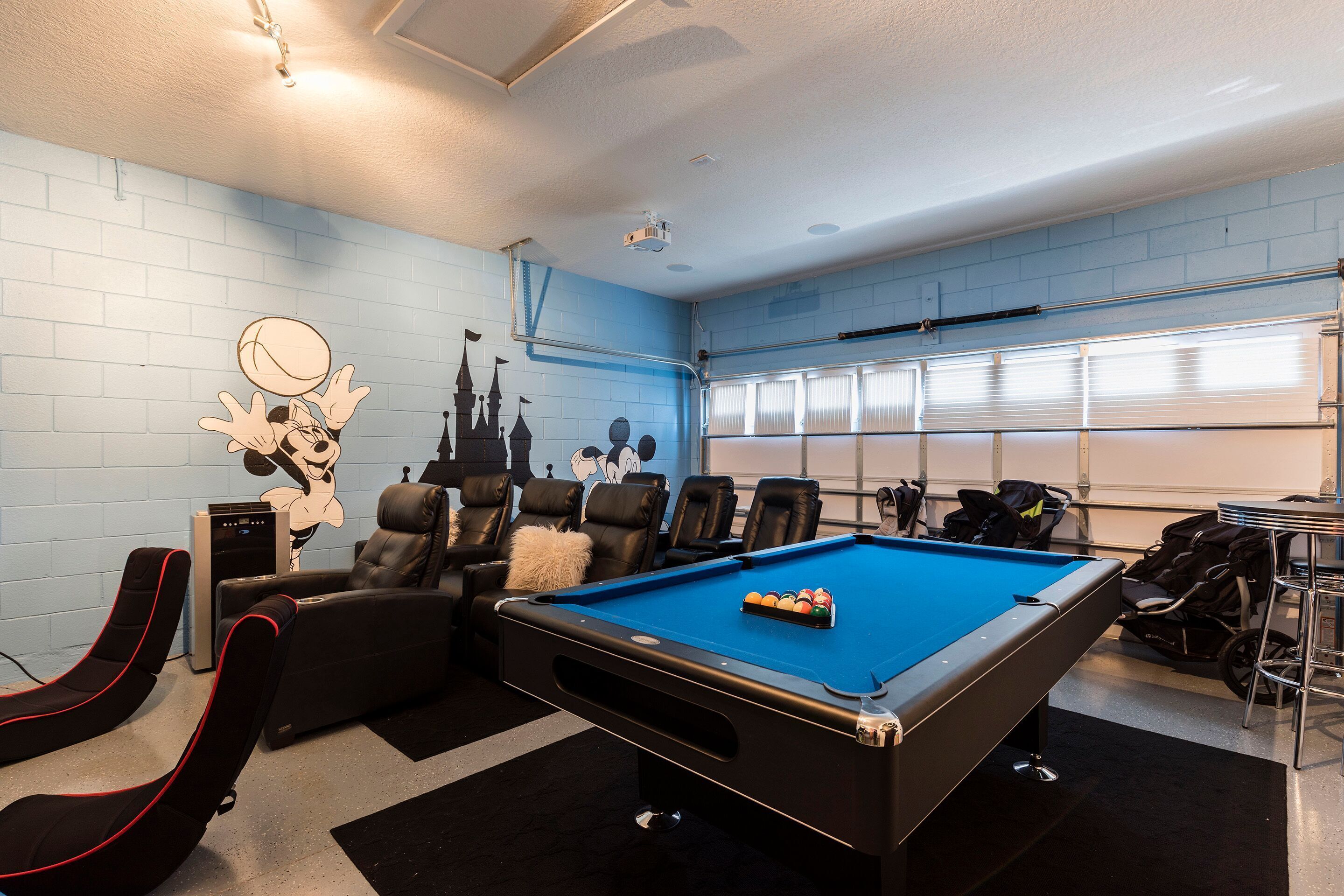 A pool table in a room with a blue brick wall, a projector, and a seating area with leather chairs.