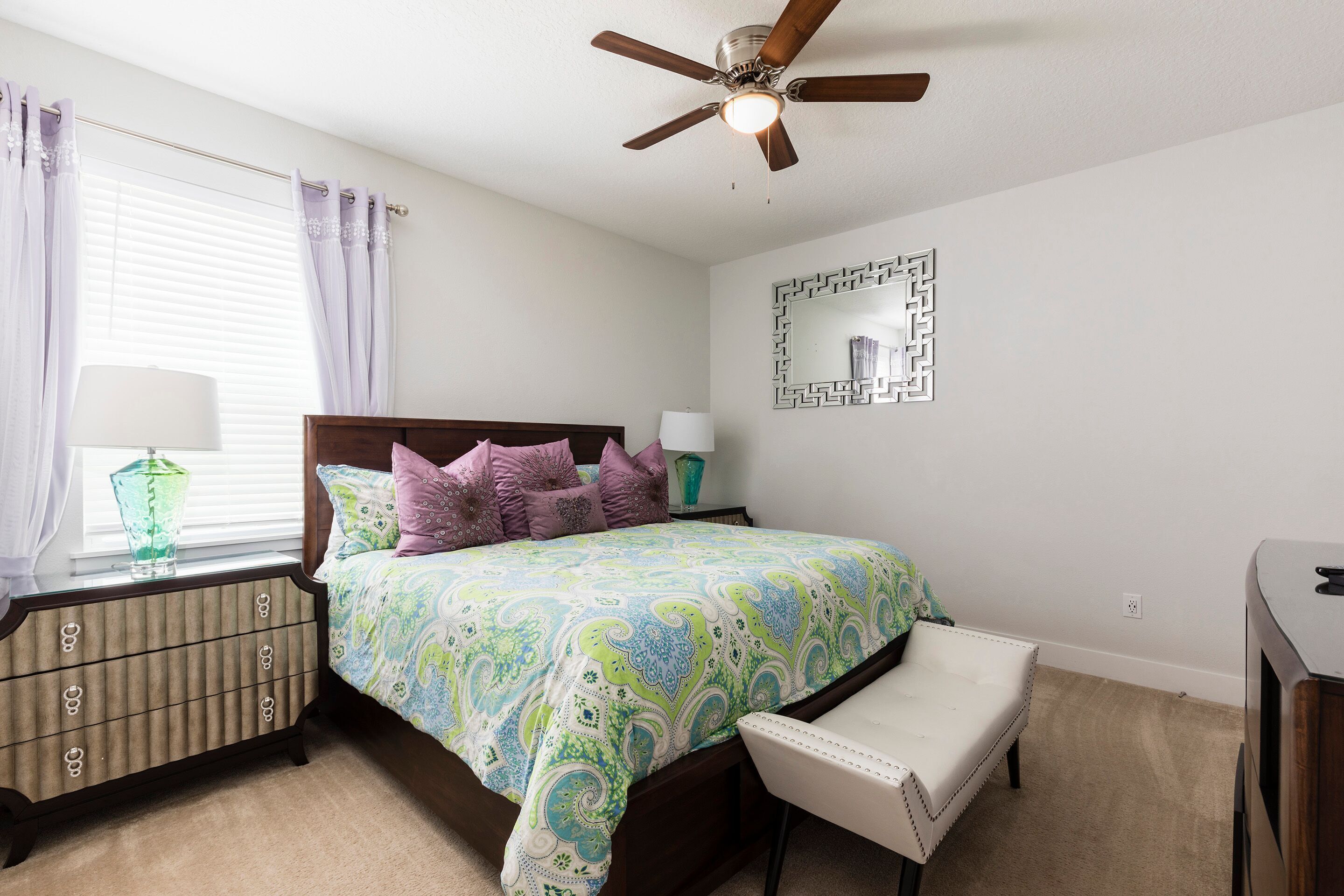 Un dormitorio con cama, mesita de noche, cómoda, silla, ventilador de techo y un espejo en la pared.
