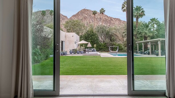 A view through a sliding glass door of a backyard with a pool, a patio area, and a mountain backdrop. - Secluded 1-Acre Mountain View Retreat - great location in La Quinta  #112390 6BR (La Quinta)