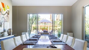 A modern dining room with a long table, white chairs, and a view of an outdoor patio with a pergola and umbrella. - Secluded 1-Acre Mountain View Retreat - great location in La Quinta  #112390 6BR (La Quinta)