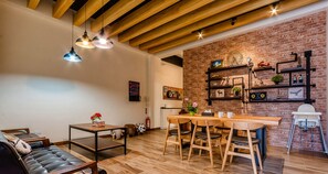 Una sala de estar acogedora con un sofá, una mesa de centro y un comedor con una mesa y sillas de madera. Se ve una pared de ladrillo con estantes y adornos al fondo.