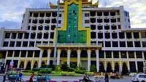 Un edificio grande de varios pisos con un elemento vertical verde y amarillo en el centro, rodeado por un estacionamiento con varios vehículos.