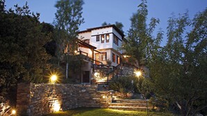 A two-story house with a stone staircase leading to the entrance, surrounded by greenery and illuminated by outdoor lighting. - PELION HOMES | Villa SELINI large traditional villa w/ pool & breathtaking view (Agios Georgios Nilias)