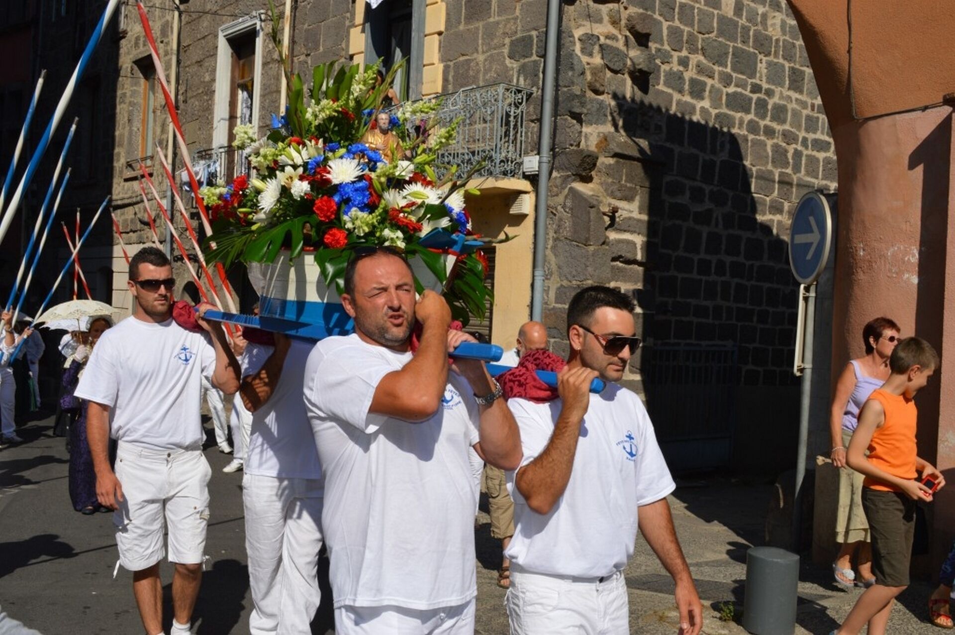 A group of people carrying a floral arrangement on a pole, walking on a street with stone buildings.