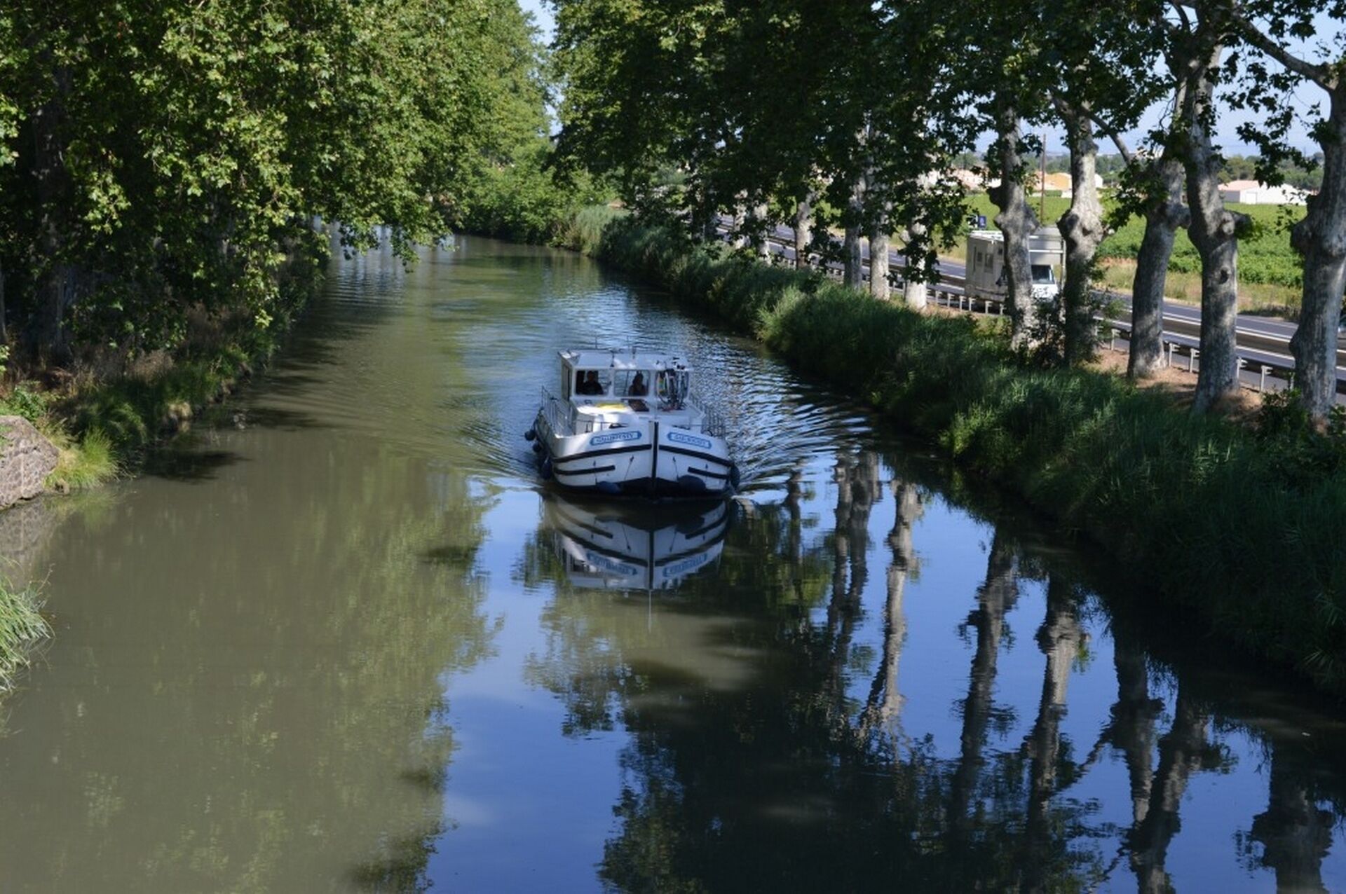 Ein schmaler Wasserweg mit einem Boot, umgeben von Bäumen und einer Straße auf der rechten Seite.
