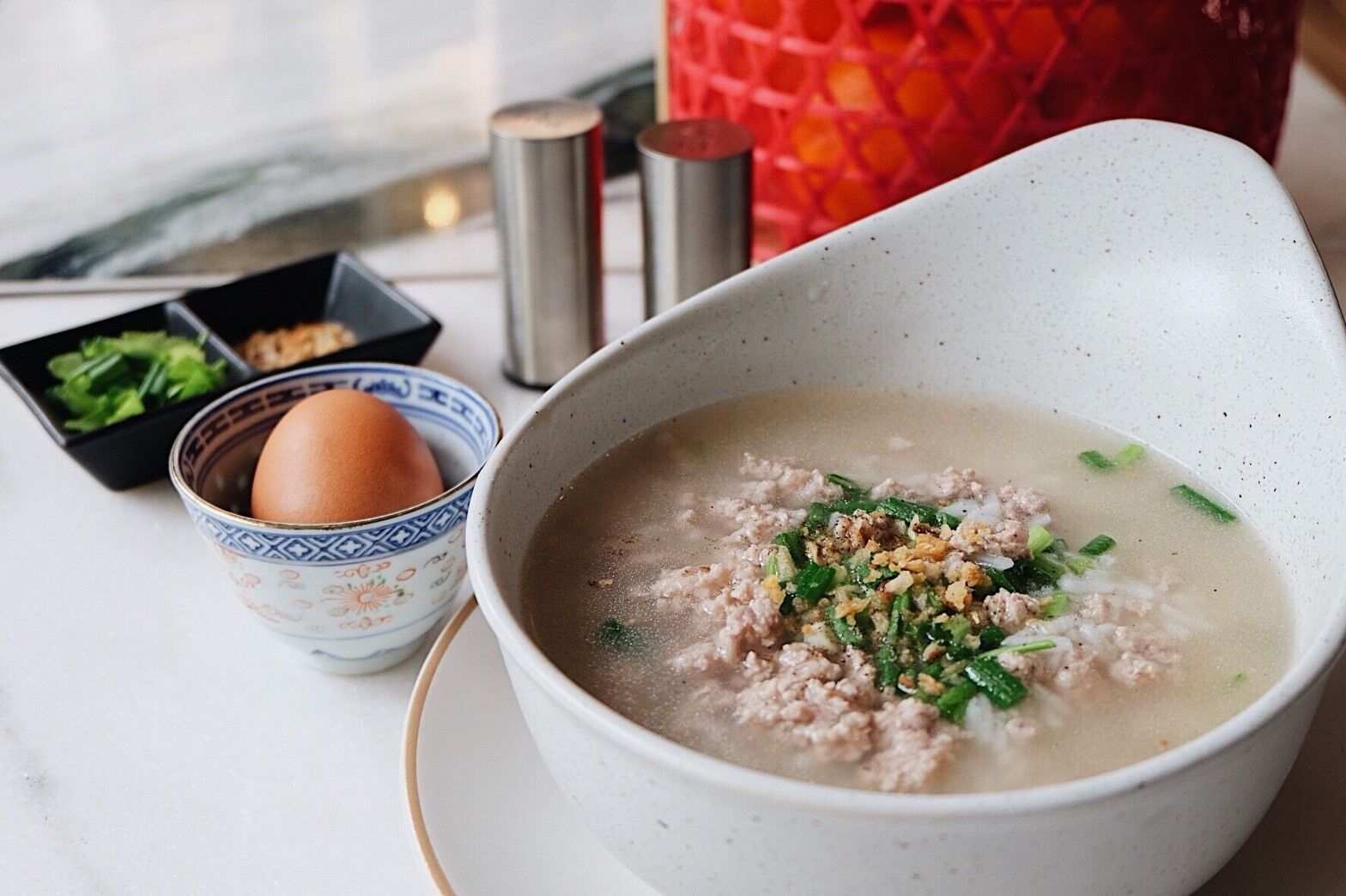A bowl of soup with ground meat and green onions, a cup with an egg, and a small bowl with green vegetables on a table.