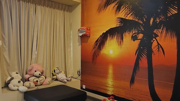 Un salon avec une grande fresque murale représentant un coucher de soleil et des palmiers, un canapé noir, une table basse en verre et une petite plante.