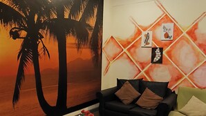 Un salon avec une grande fresque murale représentant des palmiers et un coucher de soleil, un canapé en cuir noir, une table basse en verre avec des magazines et un tapis à motifs.