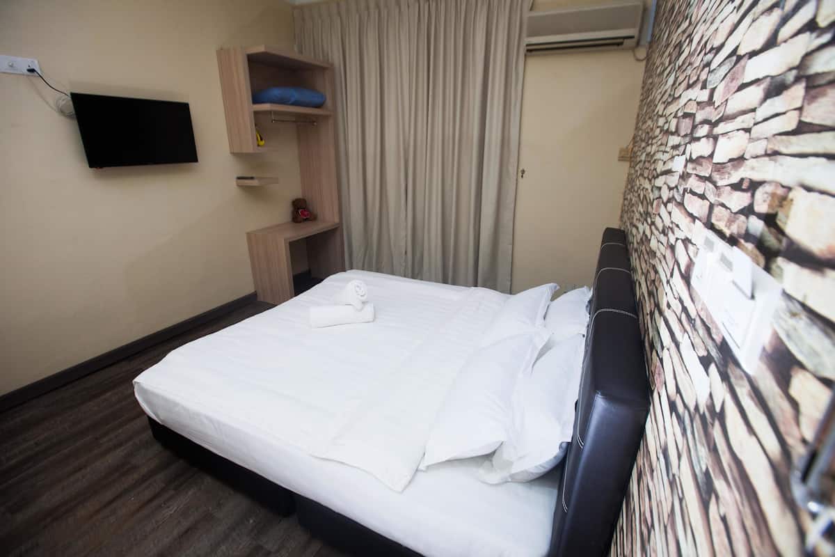 A neatly made bed with white linens, a flat-screen TV mounted on the wall, a wooden shelf with a blue cushion, and a stone-patterned wall.