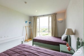 A bedroom with two beds, a window with curtains, a bedside table with a lamp, and a view of greenery outside.