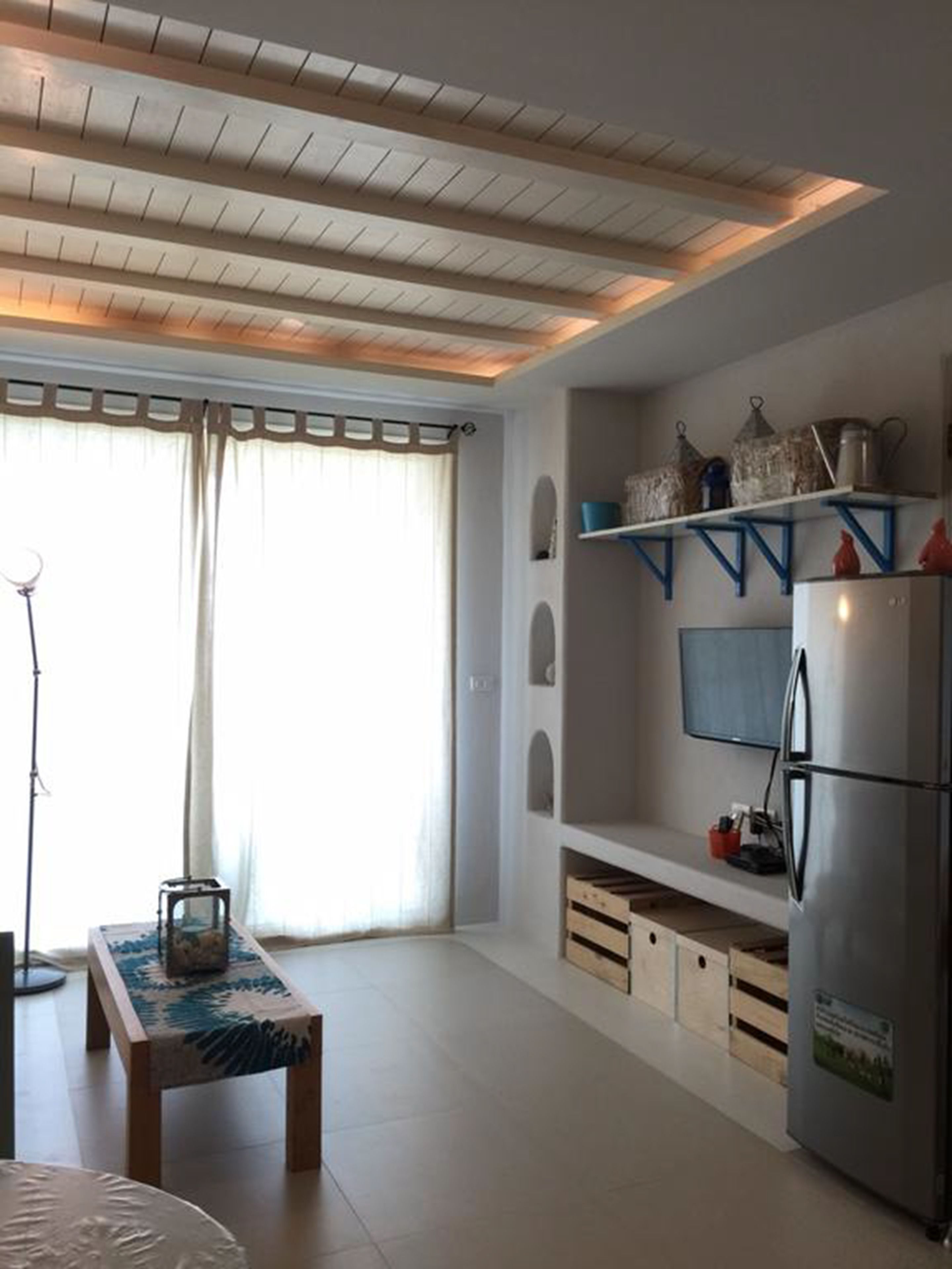 A modern kitchen with wooden ceiling beams, a built-in refrigerator, and a window with sheer curtains.