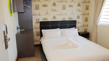 A neatly made bed with white linens and a rolled towel, a dark headboard, a small bedside table with a bottle, and a wall with a travel-themed wallpaper.