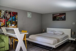 Quarto de hotel moderno com cama, escrivaninha e cadeira. Há um cartaz colorido na parede e uma pintura acima da cama.