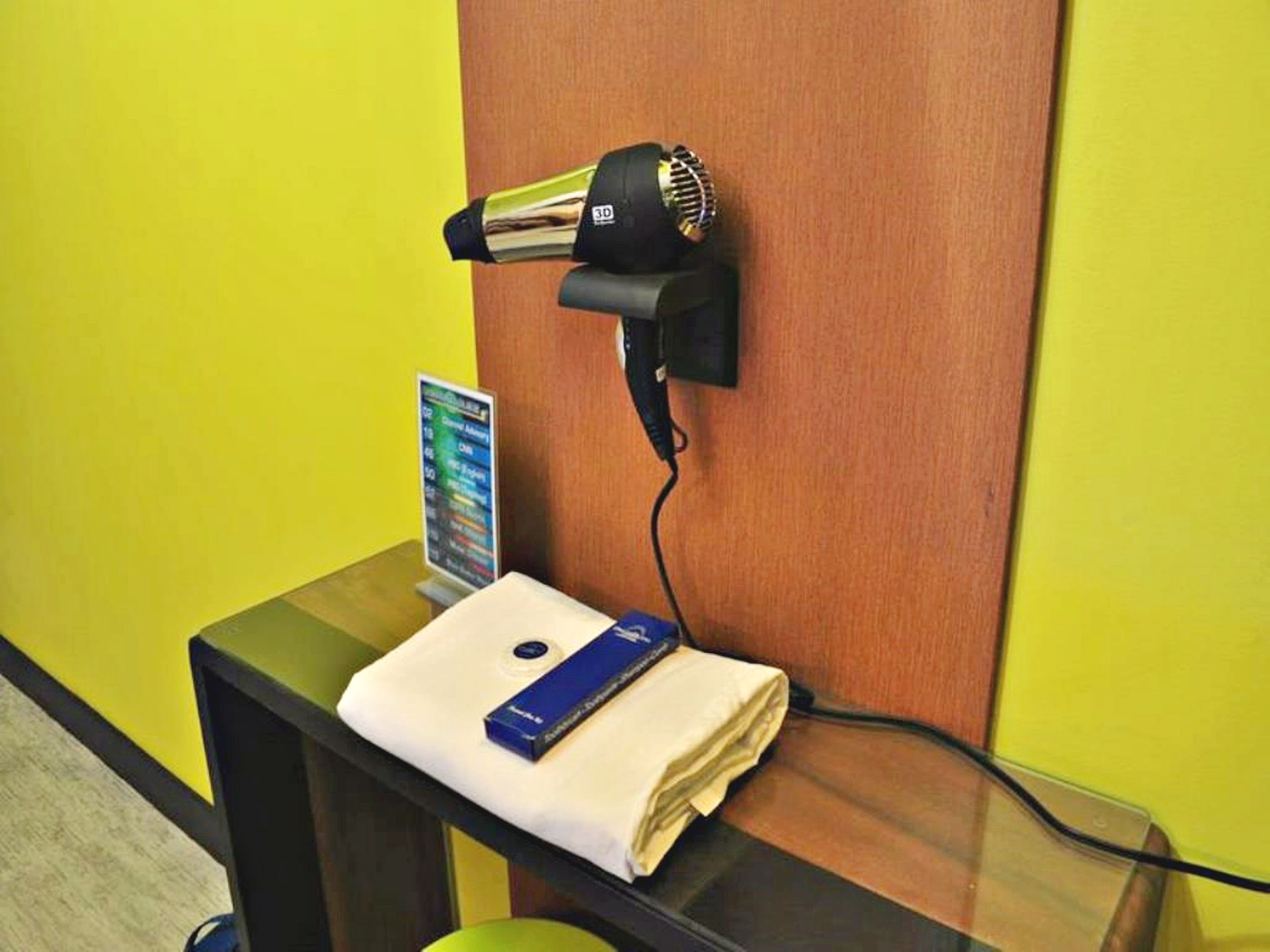 Ein an der Wand befestigter Haartrockner, ein kleiner Tisch mit einem gefalteten Handtuch und einer blauen Kiste sowie ein Schild mit einer Auflistung der Annehmlichkeiten.