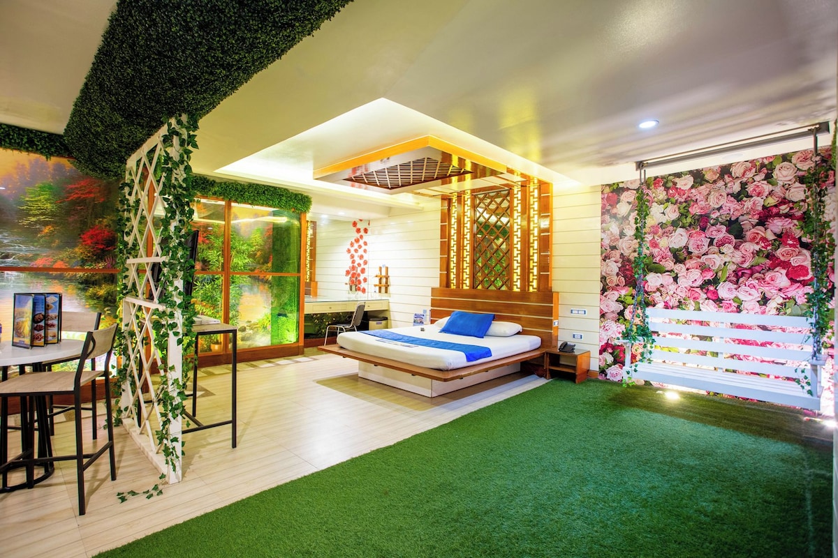 A modern hotel room with a bed, a desk, and a view of a garden through a glass wall.