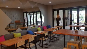 Un café moderno con mesas y sillas de madera, un sofá gris con cojines coloridos y grandes ventanales que dan a la calle.