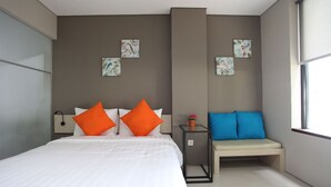 Una habitación de hotel moderna con una cama grande, dos almohadas naranjas, una mesita de noche con lámpara, un banco con cojines azules y obras de arte con motivos florales en las paredes.