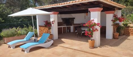 A covered outdoor seating area with lounge chairs and a table, surrounded by potted plants and a barbecue grill.