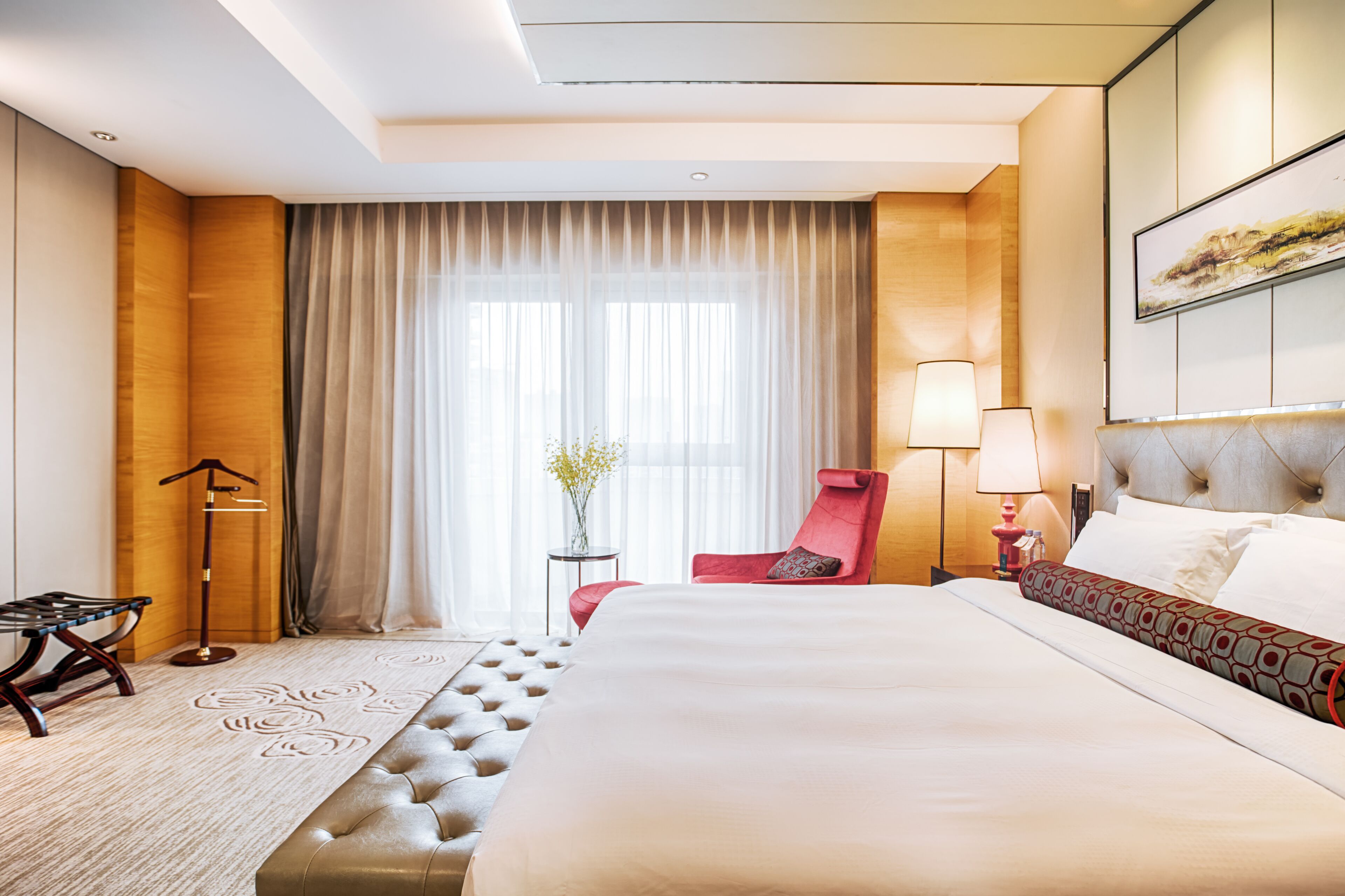 Una habitación de hotel con una cama grande, un sillón rojo, una lámpara de pie y una mesita con un florero.