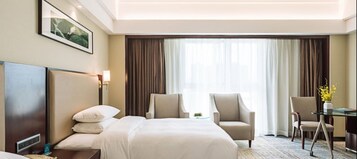 Habitación de hotel con una cama, dos sillones, un escritorio con un florero y un ventanal con cortinas.