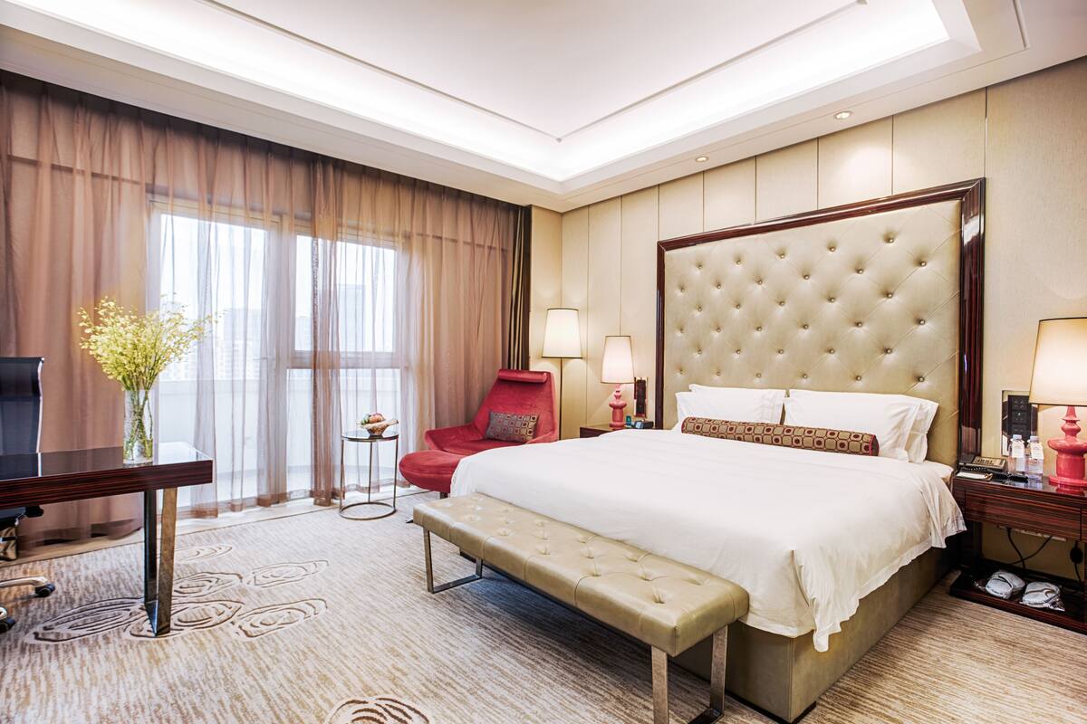 A hotel room with a large bed, a desk, a chair, a bench, and a TV.