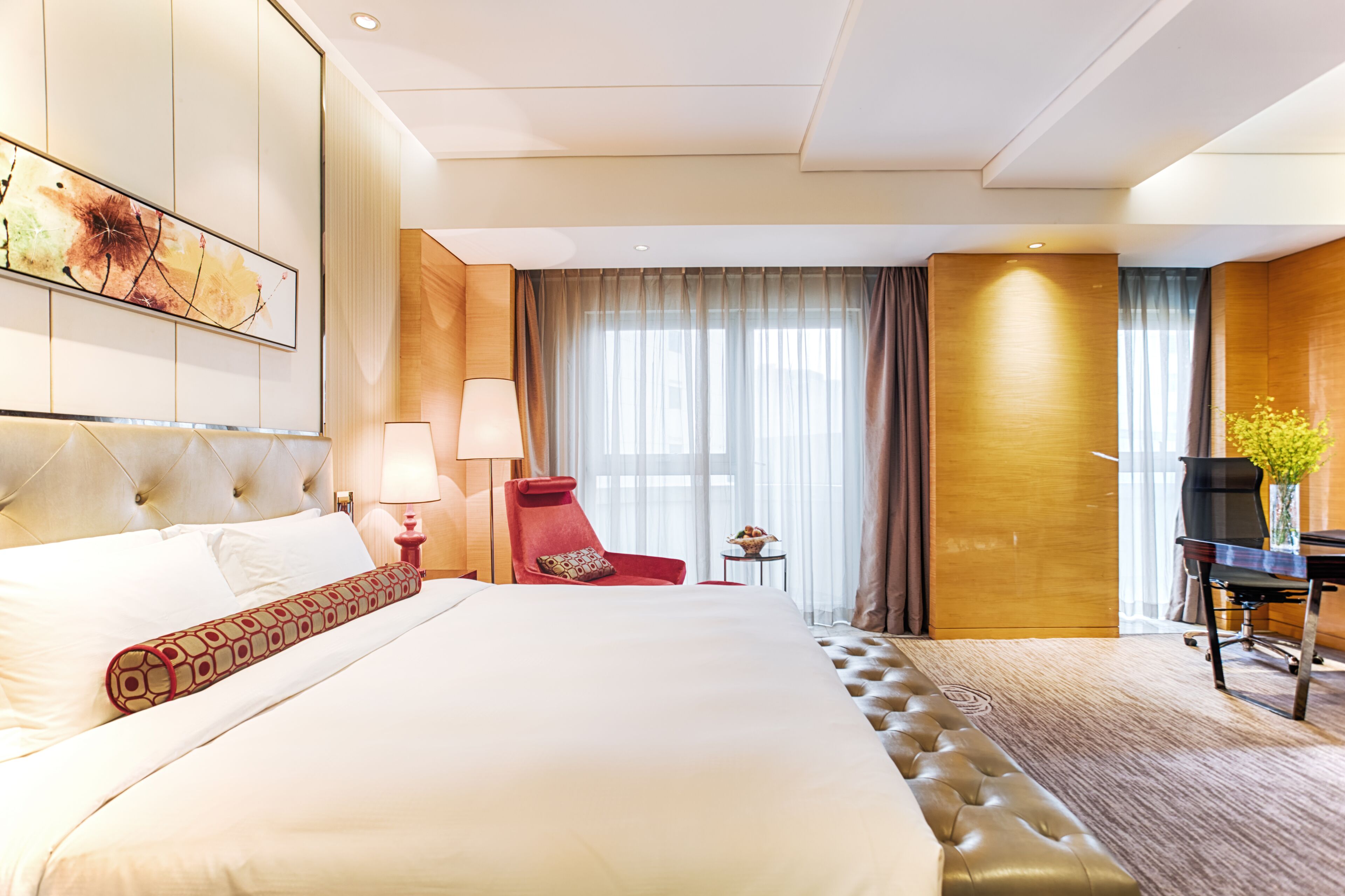 Habitación de hotel con una cama grande, un sillón rojo, un escritorio con una silla, una lámpara y un cuadro en la pared.