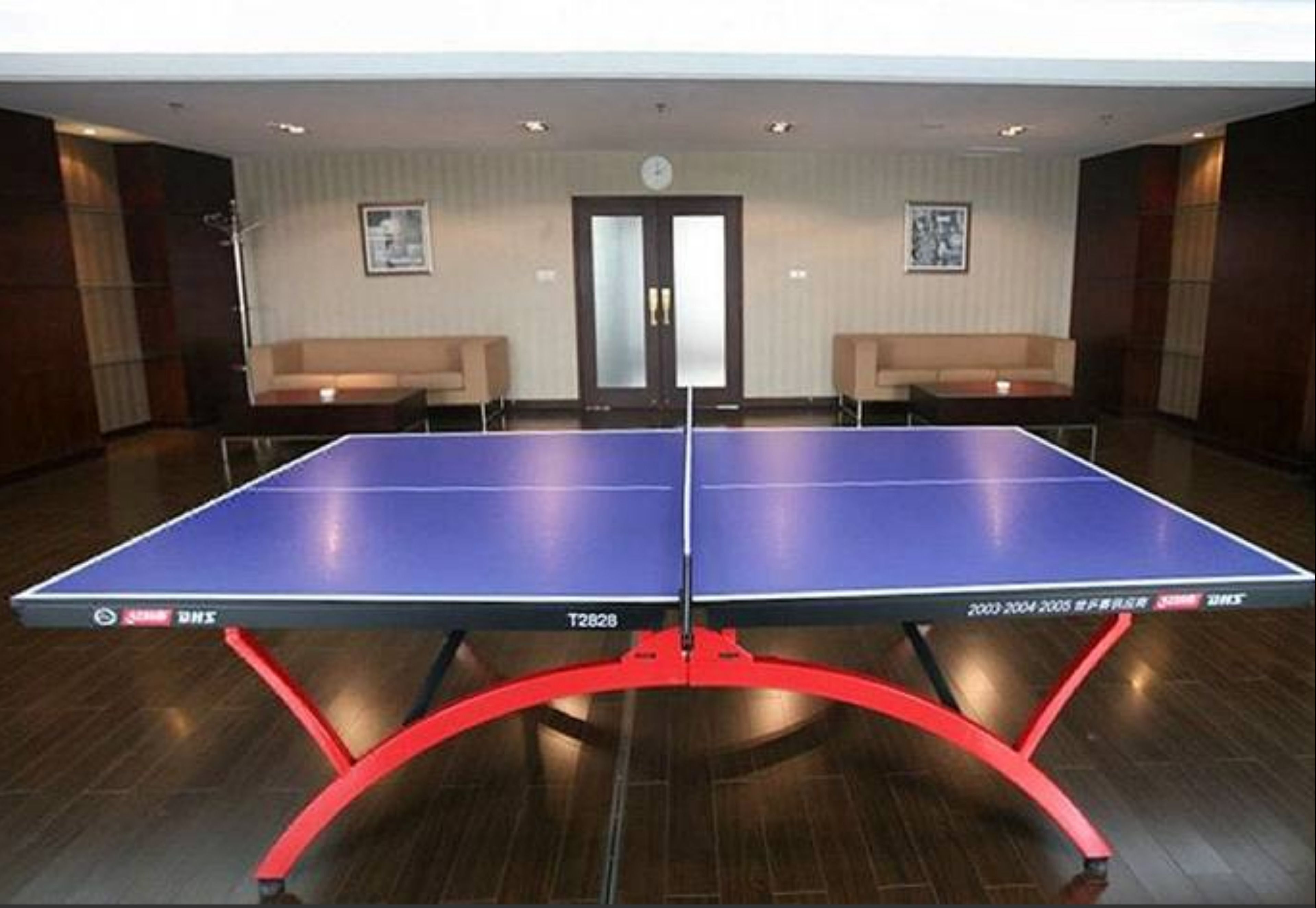 A ping pong table in a hotel room with a wooden floor, two sofas, and a door with glass panels.