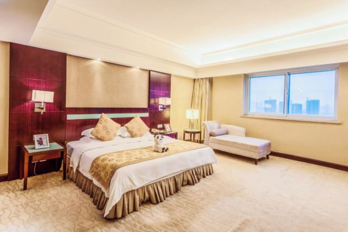 A hotel room with a large bed, a sofa, a small table, and a window with a city view.