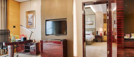 Et hotelværelse med et fladskærms-tv, et skrivebord med lampe, en kommode og et tæppe på gulvet.