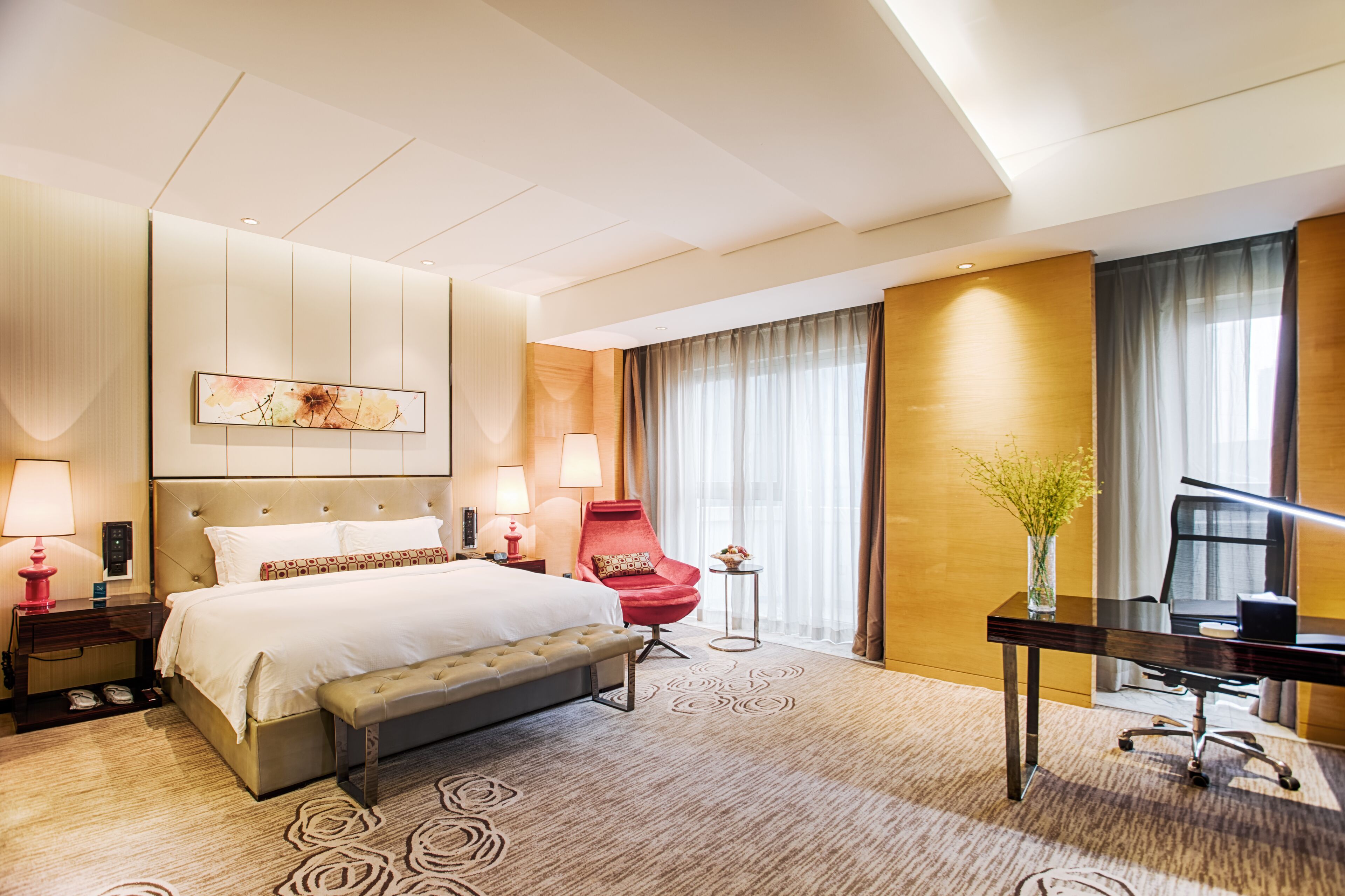A hotel room with a large bed, a desk, a red armchair, and a small table with a vase of flowers.