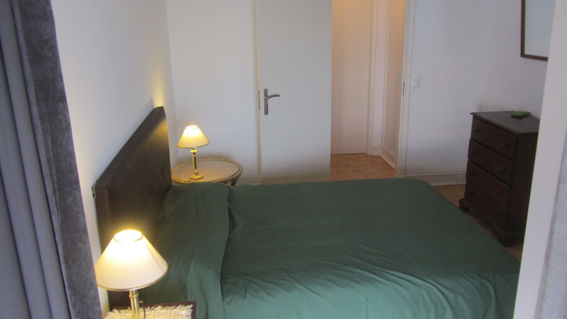 A small, simple room with a bed, two bedside lamps, a door, and a dresser.