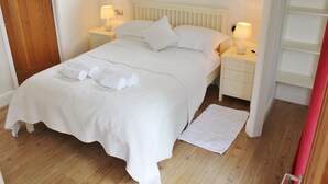 En pænt redt seng med hvide sengetøj og håndklæder, et natbord med lampe, og trægulv.