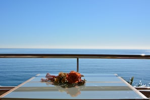 Een balkon met een glazen tafel en een bloemstuk, met uitzicht op een rustige zee.