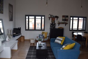 A modern living room with a blue sofa, a coffee table, and a television. There are two windows with black frames, a dining area with a table and chairs, and a kitchen area with shelves and appliances.