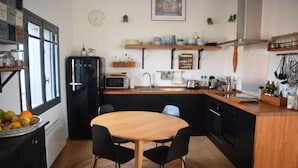 Een kleine keuken met een houten tafel en stoelen, een koelkast, een magnetron en diverse keukengerei.