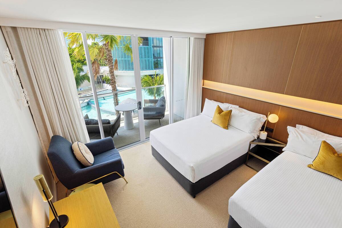 A hotel room with two beds, a sofa, a small table, and a view of a pool and palm trees.