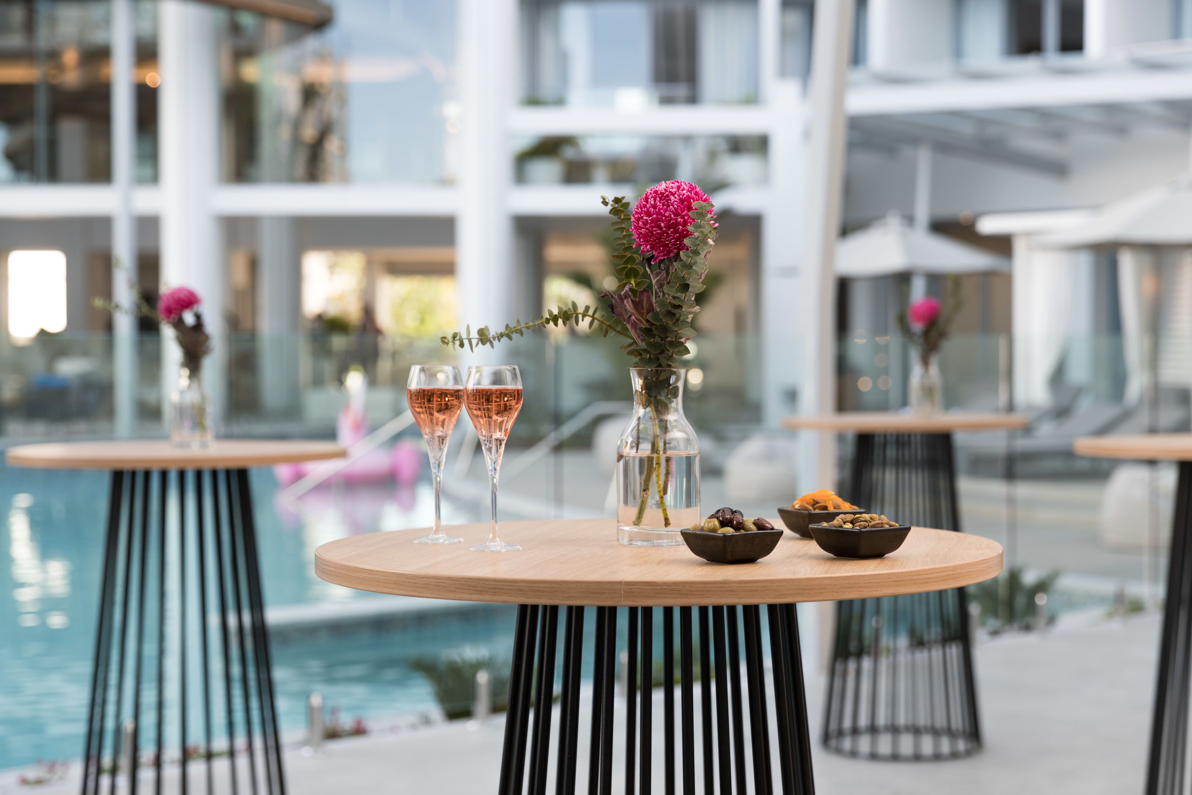Ein Tisch am Pool mit zwei Gläsern Roséwein, einer Vase mit einer rosa Blume und einer Oliven-Schale.