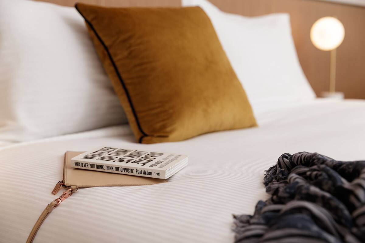 A neatly made bed with a book titled "Whatever You Think, Think the Opposite" by Paul Arden, a brown pillow, and a bedside lamp.