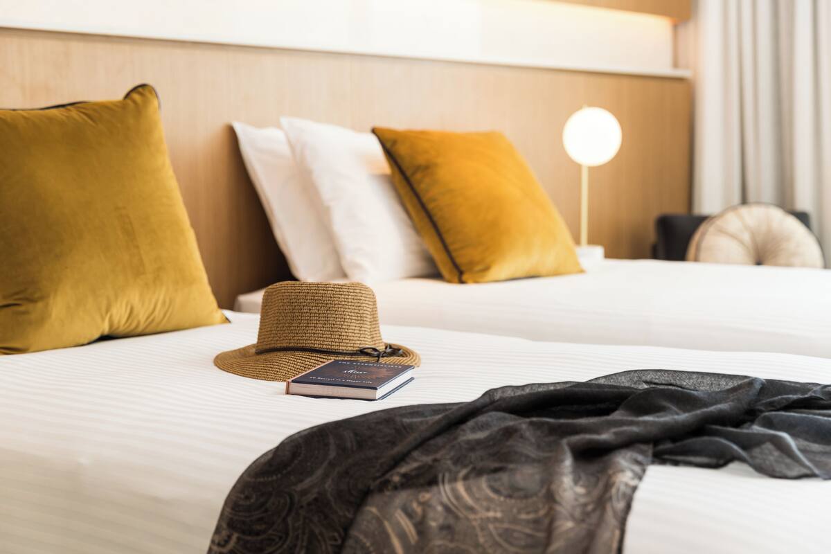 A neatly made bed with a hat and book on it, surrounded by pillows and a bedside lamp.