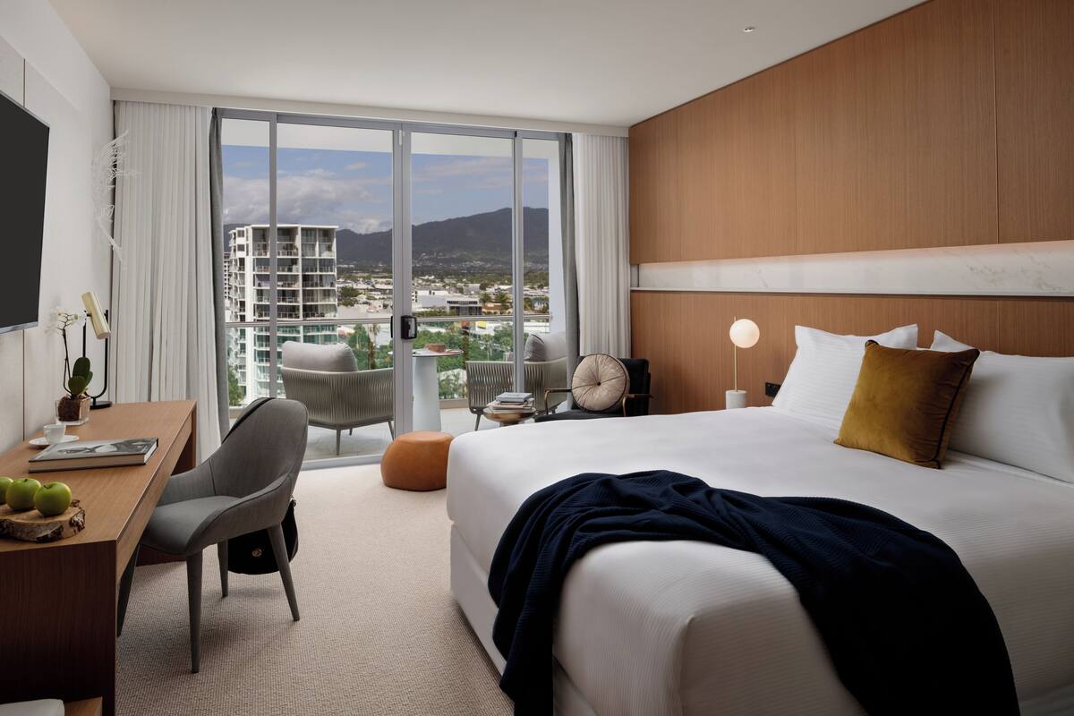 A hotel room with a large bed, a desk with a chair, a dining table with apples, and a view of the city through large windows.