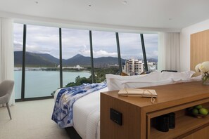 A hotel room with a large window offering a view of a body of water and mountains. There is a bed with a blue and white floral bedspread, a wooden desk with a handbag, and a vase with flowers.