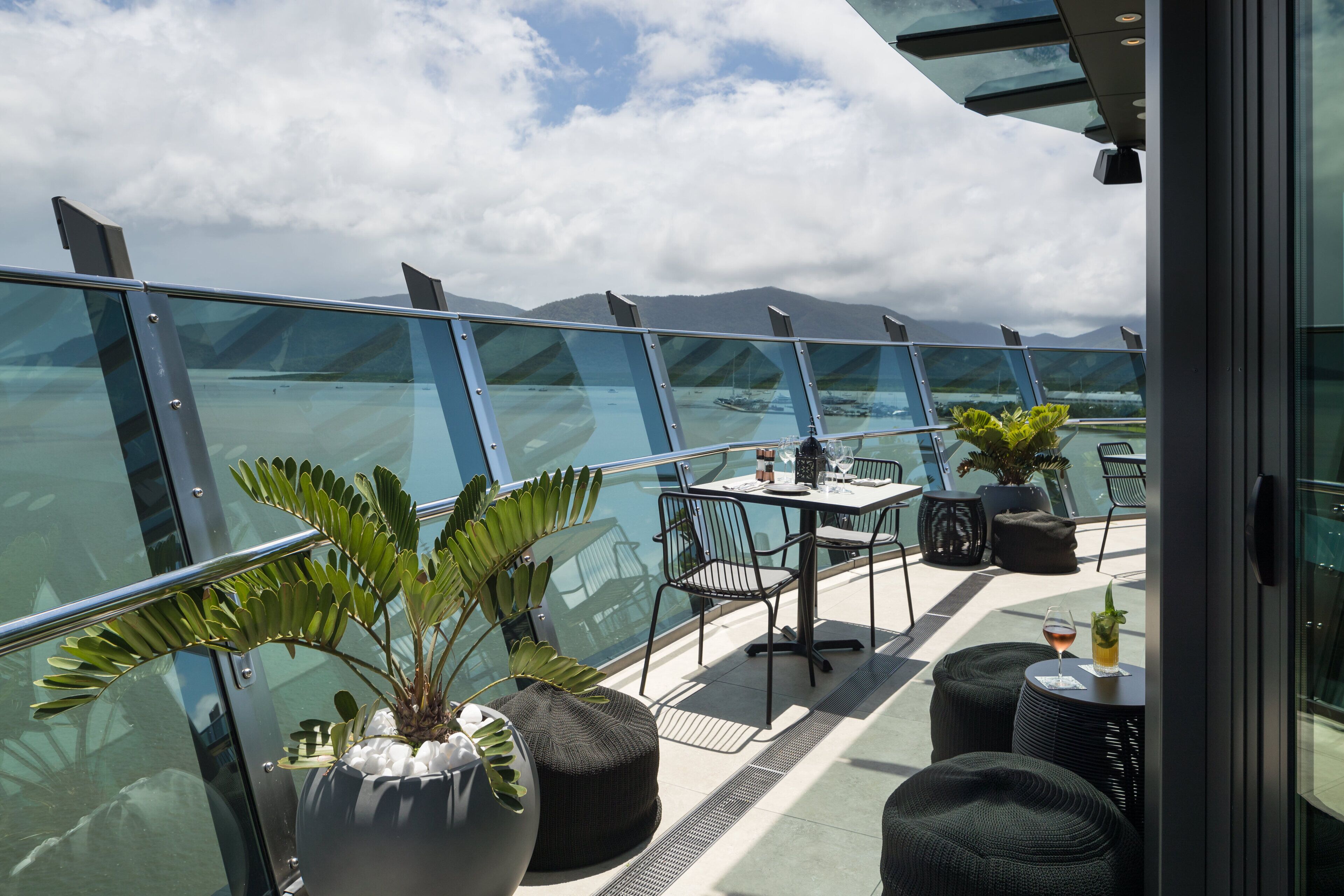 A modern outdoor seating area with glass railings, a table with chairs, and potted plants.