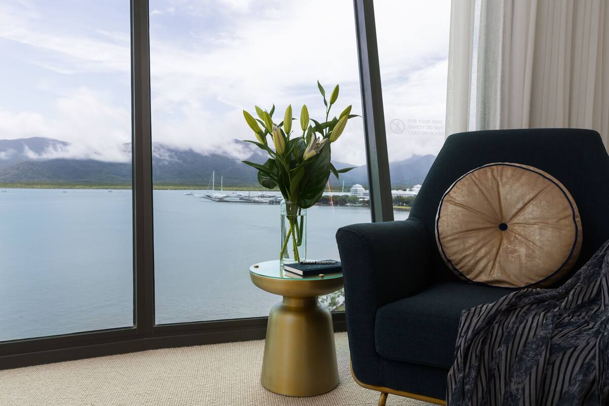 A room with a large window offering a view of a body of water and mountains. There is a round side table with a vase of flowers and a book. A dark blue armchair with a round cushion is positioned next to the window.