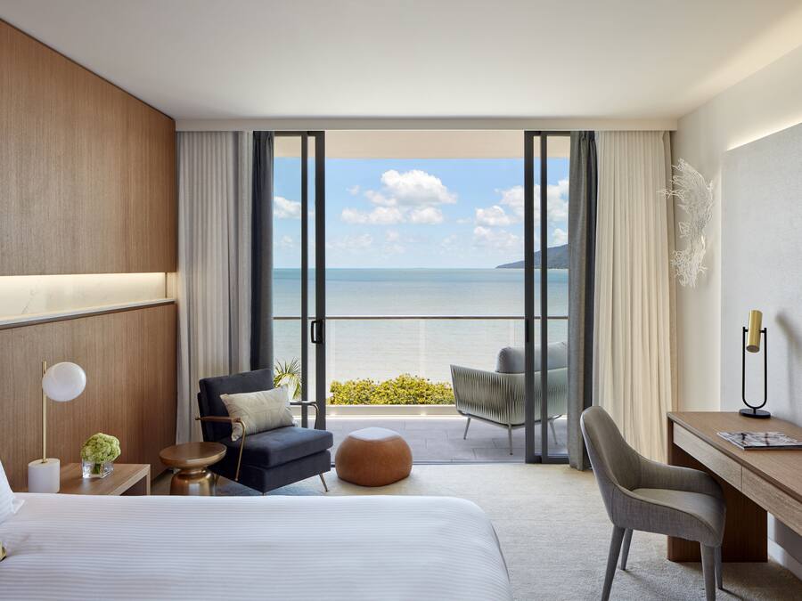 Ein Hotelzimmer mit einem Bett, einem Schreibtisch, einem Sessel, einem kleinen Tisch und Meerblick durch große Fenster.
