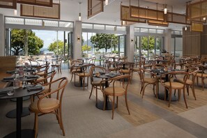 A modern restaurant with round tables and wicker chairs, large windows offering views of trees and a body of water.